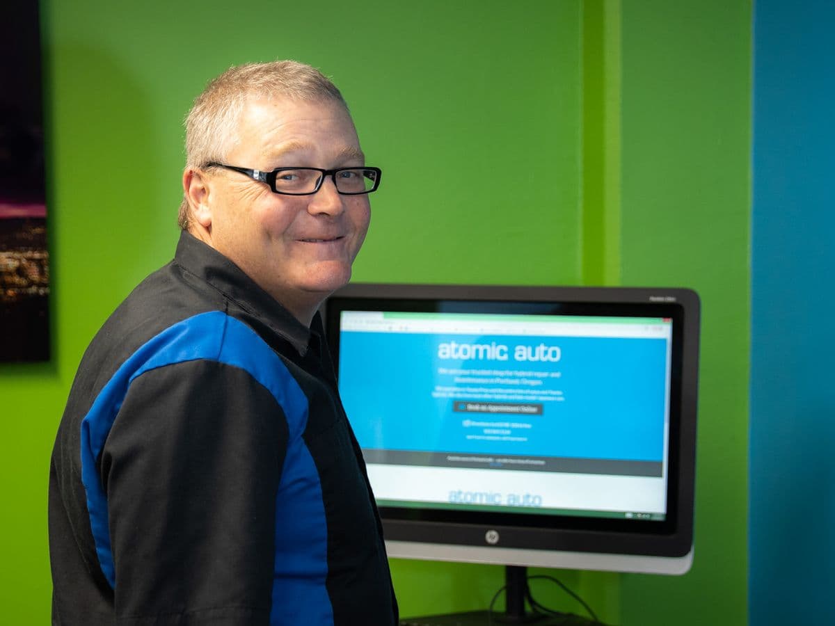 Travis Decker in the blue and black Atomic Auto shirt, smiling over his shoulder at the front-desk computer