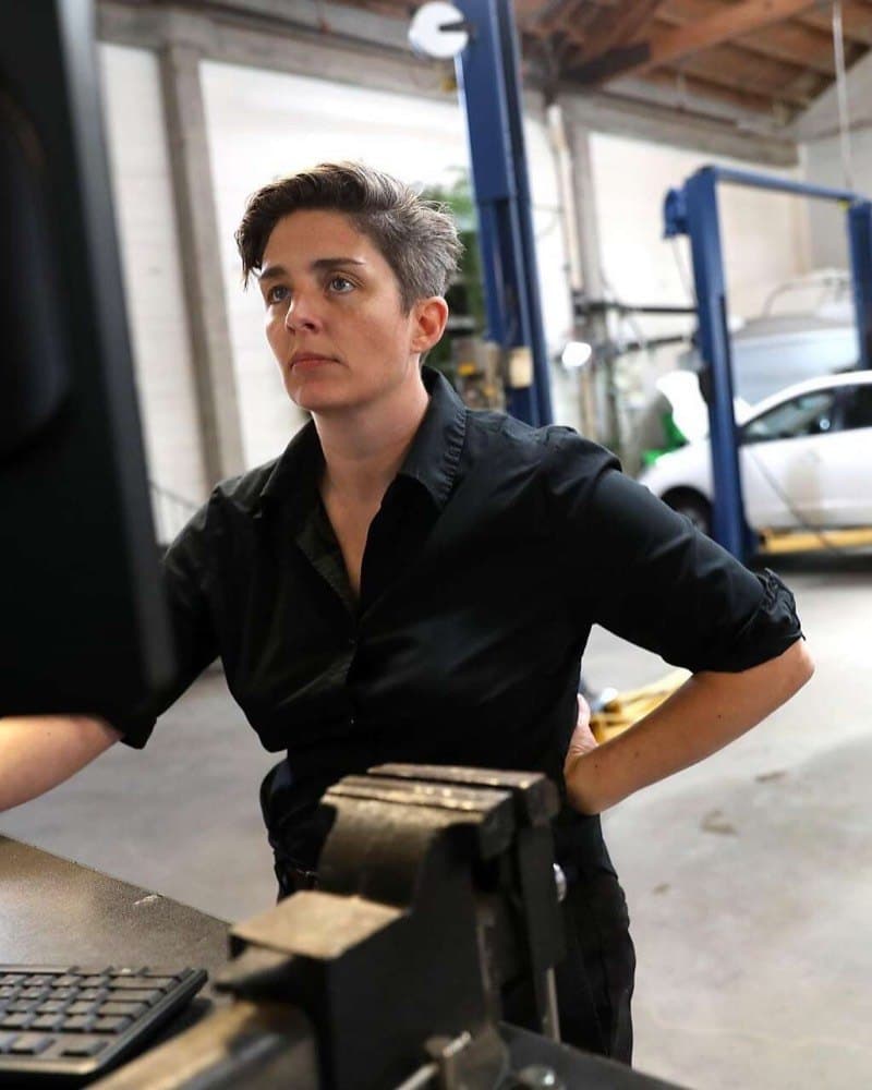 Carolyn Coquillette at a workbench in the shop, a car up on a blue lift behind her